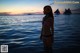 A woman in a bikini standing in the ocean at sunset.